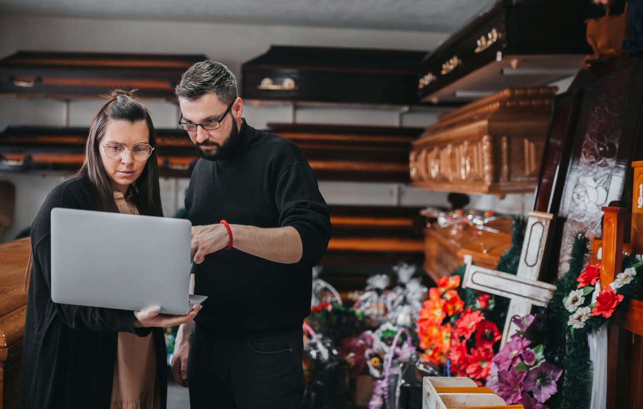 Two people stand in a funeral home, looking at a laptop. They are surrounded by coffins, flower arrangements, and funeral decor, appearing focused on the screen as they discuss something.