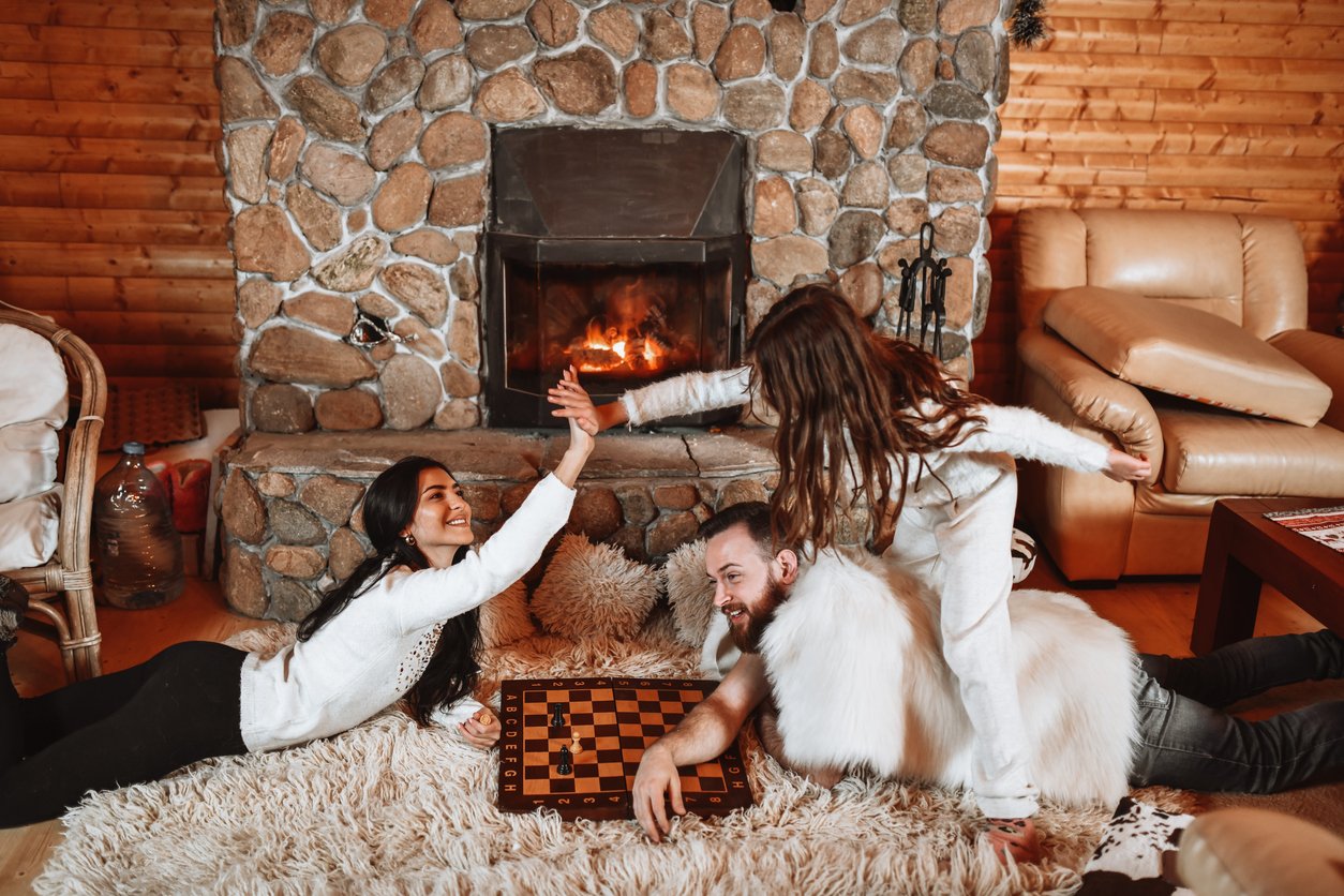 A family of three, dressed in cozy white clothes, smiles and high-fives while lying on a rug in front of a stone fireplace, with a chessboard between them in a warm, rustic living room.