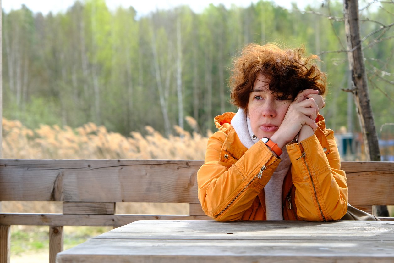 A woman with curly red hair wearing an orange jacket sits at a wooden table outdoors, resting her head on her hands and gazing thoughtfully into the distance. Trees and reeds are visible in the background.