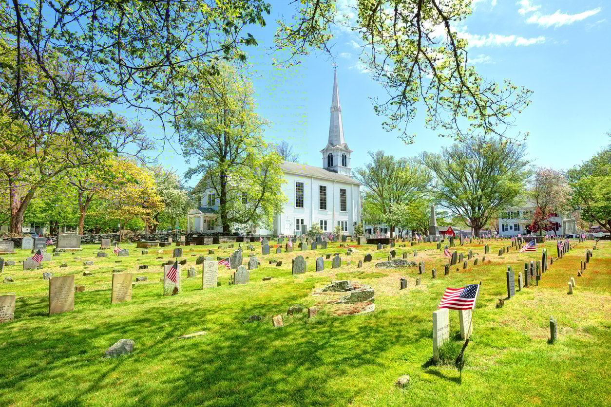 A historic cemetery with old gravestones and American flags on a bright, sunny day, with a white church and steeple in the background surrounded by leafy trees.