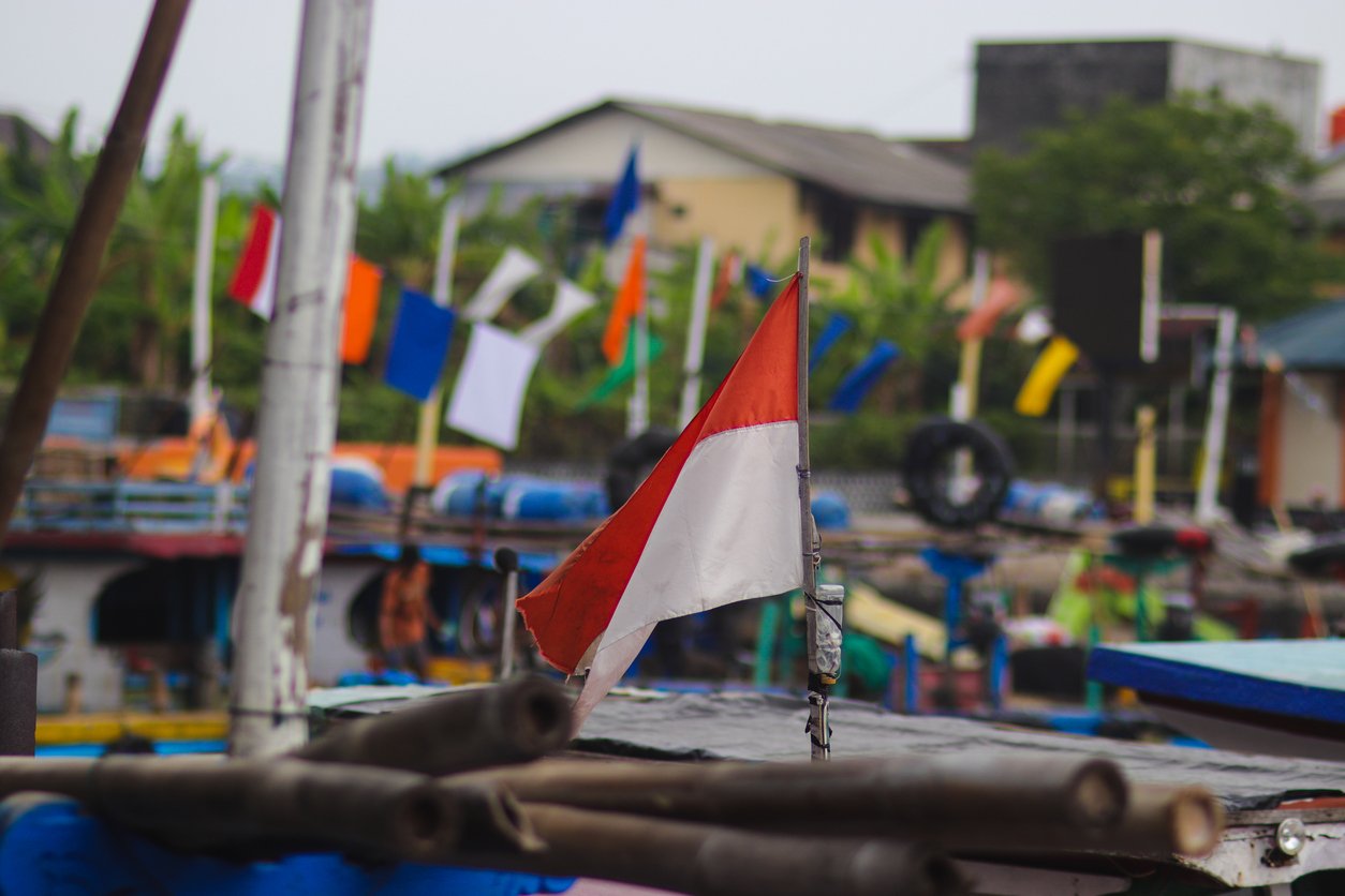 A red and white flag flutters on a boat in a busy harbor, with colorful flags and blurred buildings and greenery visible in the background.