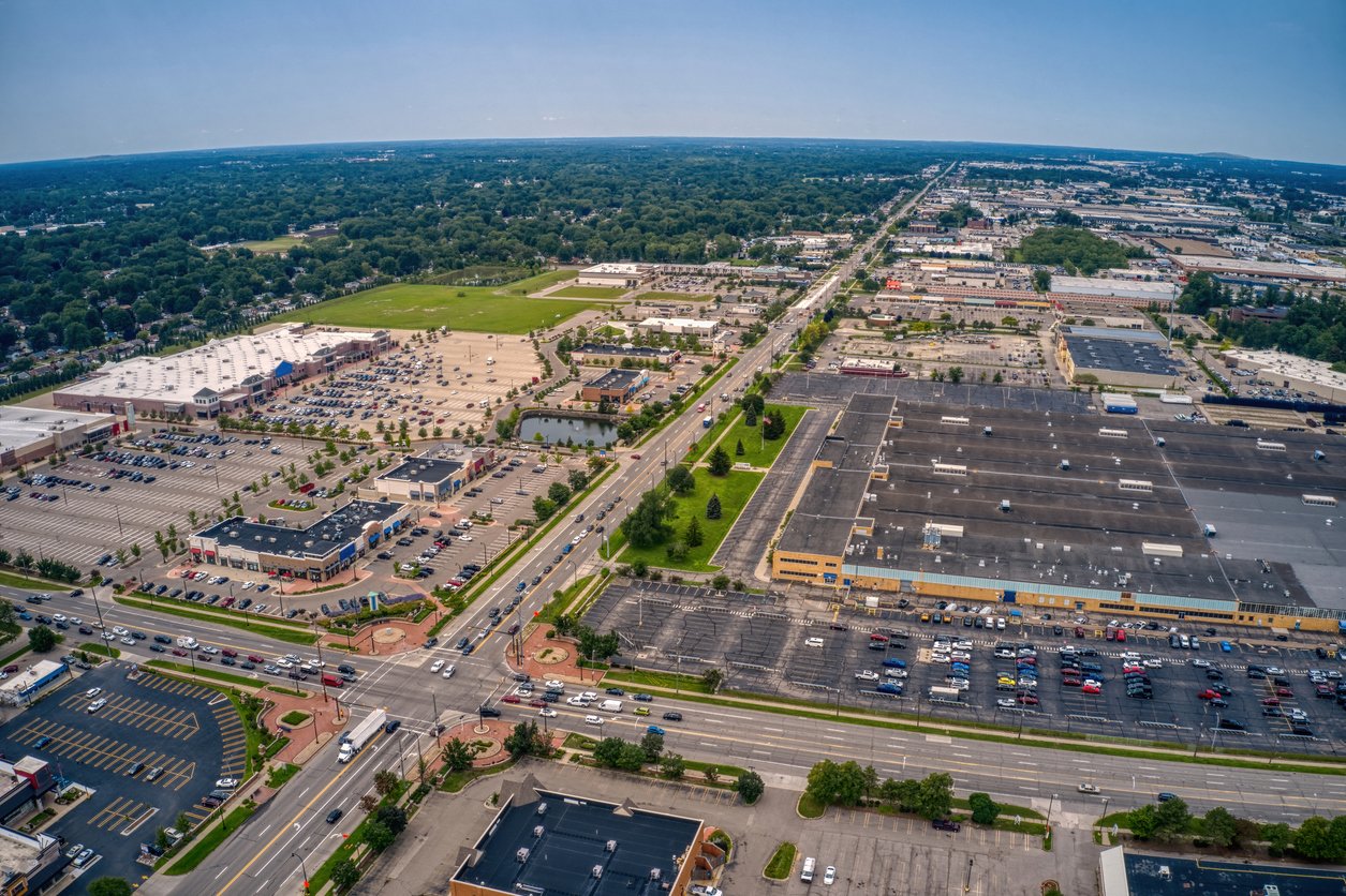 Aerial view of a suburban area with large shopping centers, parking lots, and a main road running through the middle, surrounded by green trees and distant residential neighborhoods.