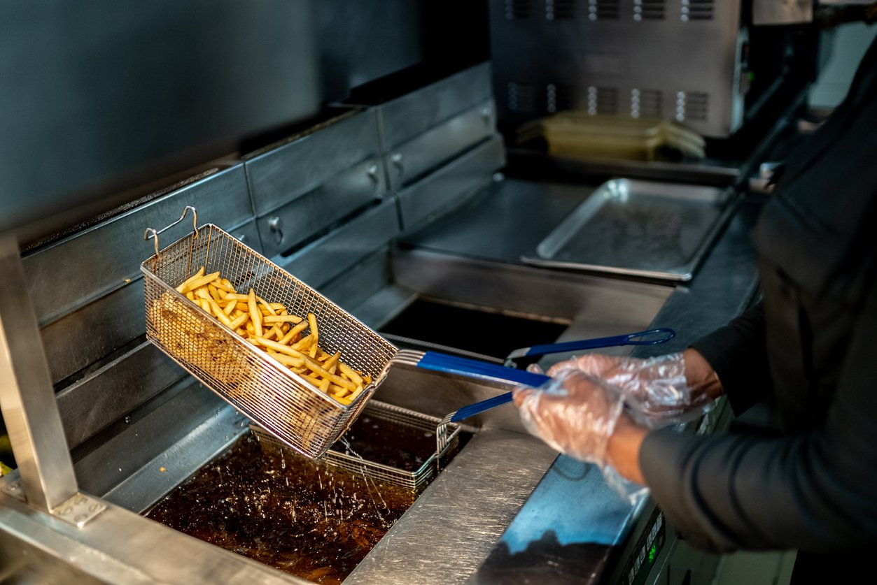 A person wearing gloves lifts a metal basket of freshly cooked French fries from a deep fryer in a commercial kitchen.