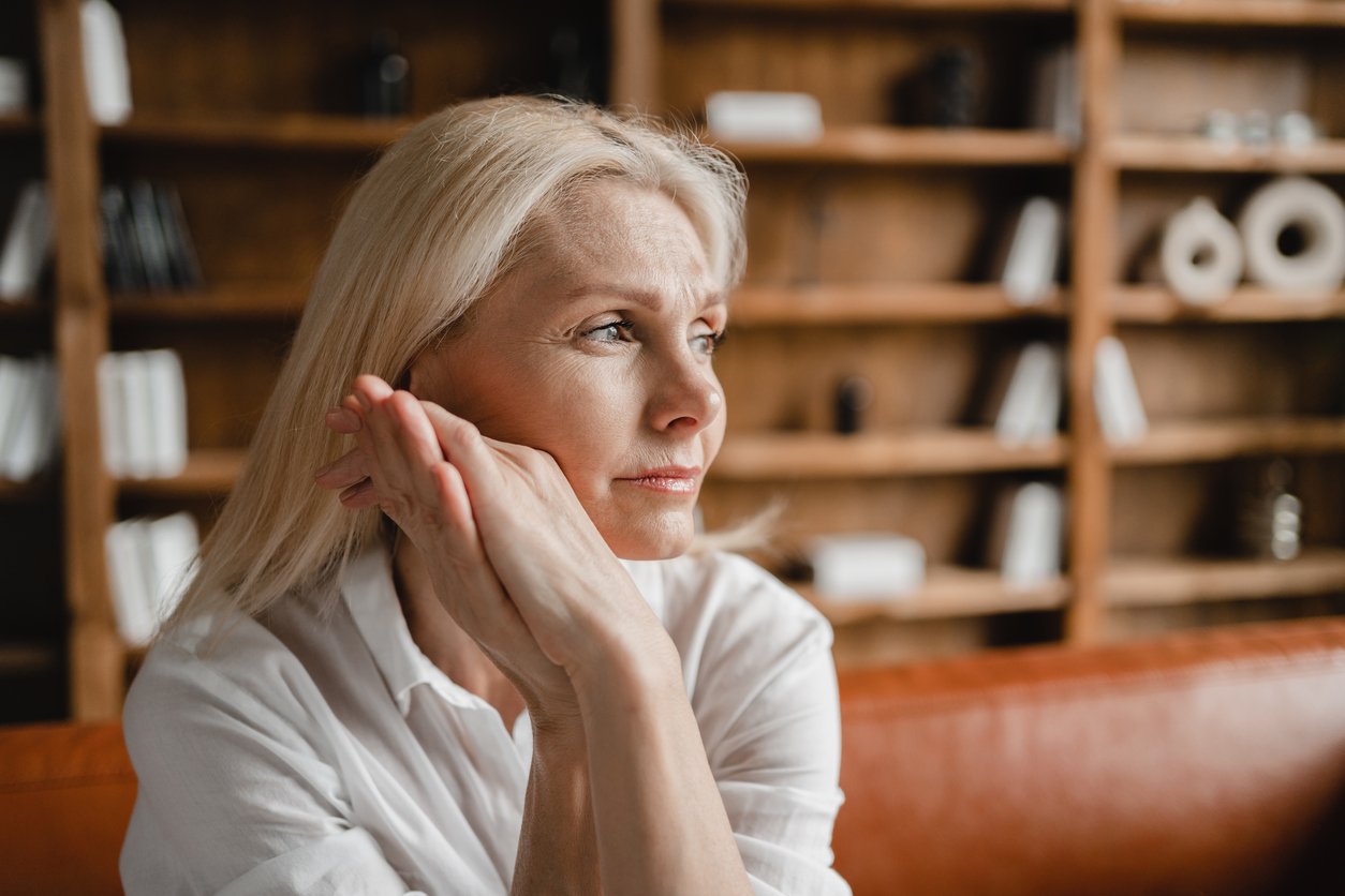 A woman with blonde hair sits indoors, resting her hands on her cheek and looking thoughtfully to the side. She is wearing a white shirt, and there are shelves with books and decor in the background.