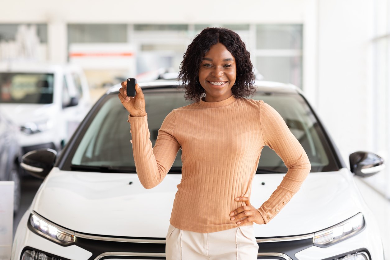 A smiling woman stands in front of a white car, holding up a car key, suggesting she just purchased or received the vehicle.
