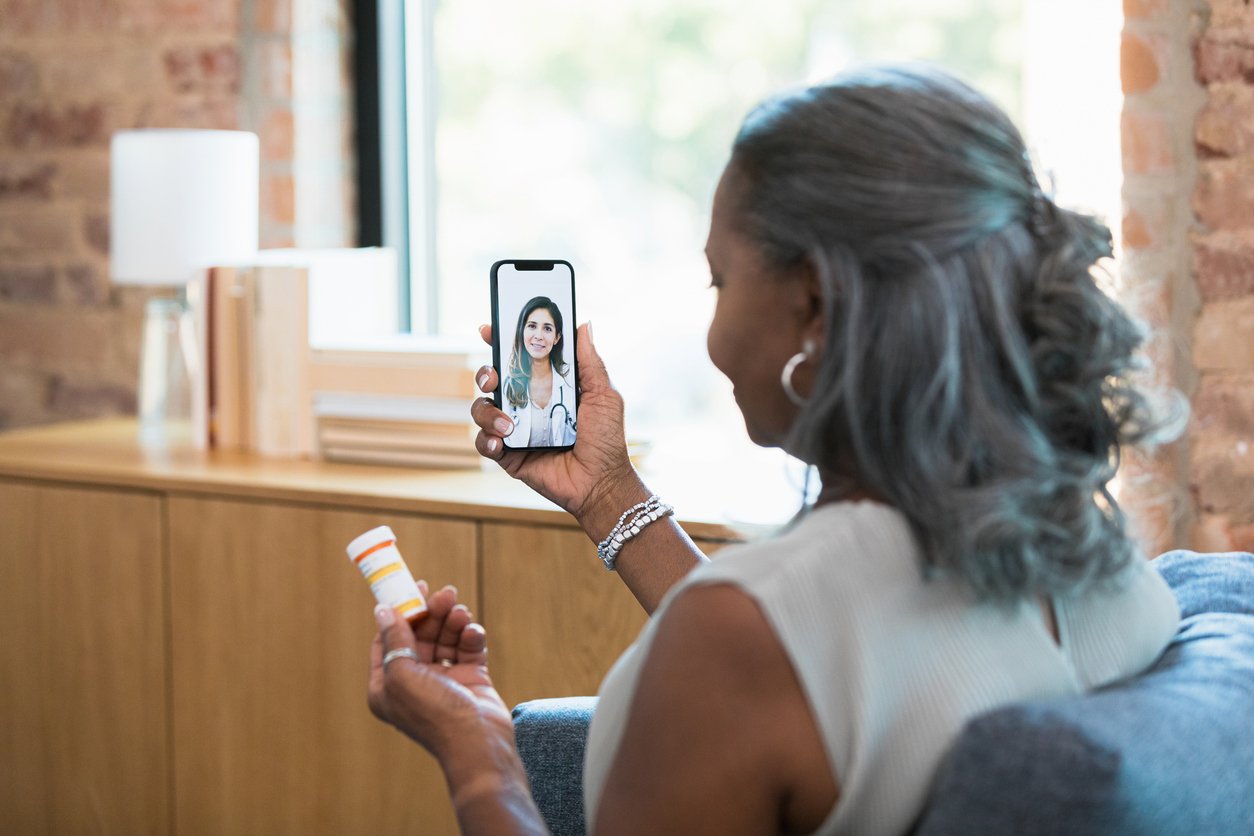 An older woman sits on a couch holding a prescription bottle and video calling a doctor on her smartphone, likely discussing her medication, in a bright, homey living room.