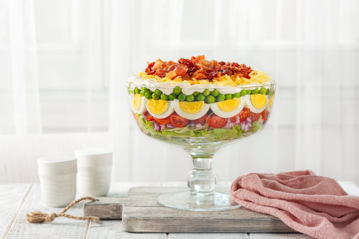 A glass trifle bowl filled with a layered salad, including lettuce, tomatoes, red onions, peas, sliced hard-boiled eggs, shredded cheese, and crispy bacon. The bowl sits on a wooden board near cups and a pink cloth.