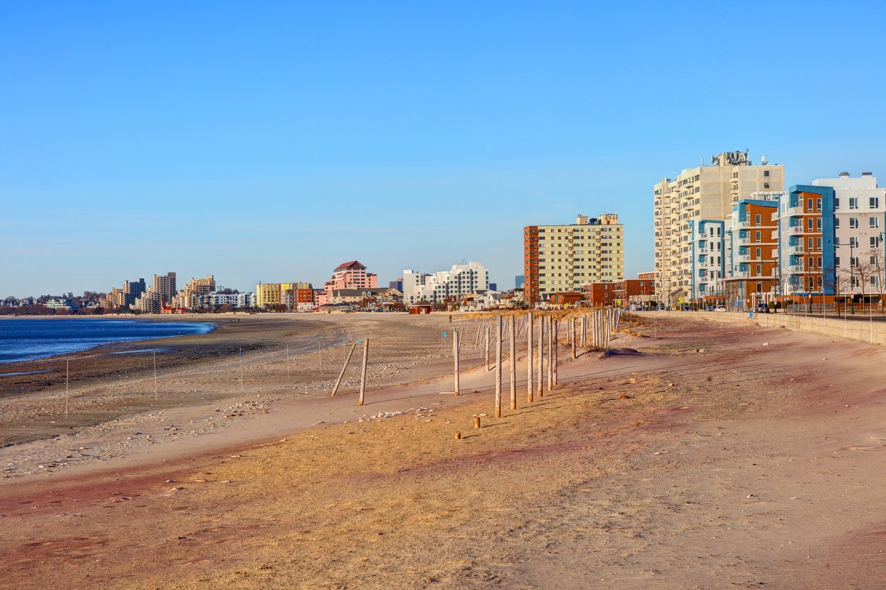 A sandy beach with scattered fencing, calm waves, and several mid-rise apartment buildings and hotels in the background under a clear blue sky.