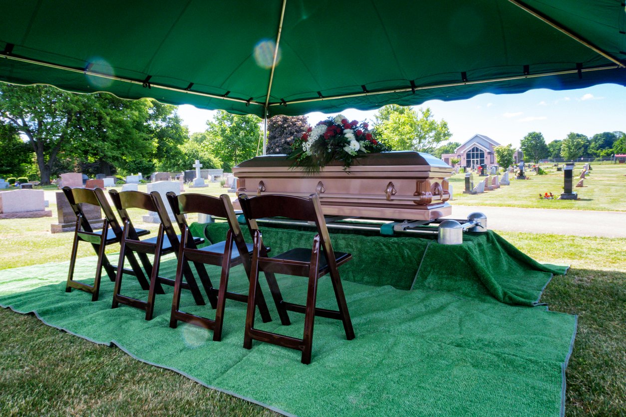 A casket adorned with flowers sits under a green tent at a cemetery, surrounded by empty chairs on green rugs, with gravestones and trees visible in the background on a bright day.