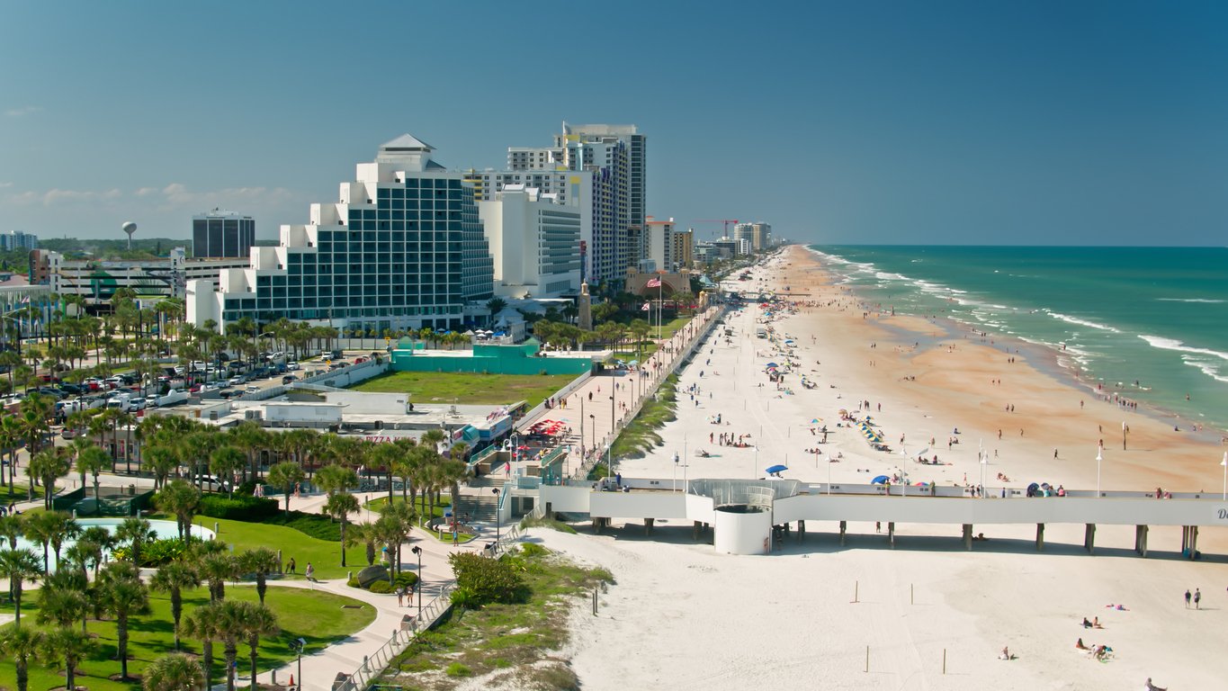 Aerial view of a coastal city with tall hotels and buildings lining a sandy beach. People walk and relax along the shore, and palm trees dot the green areas near the beach. The ocean is calm and blue under a clear sky.