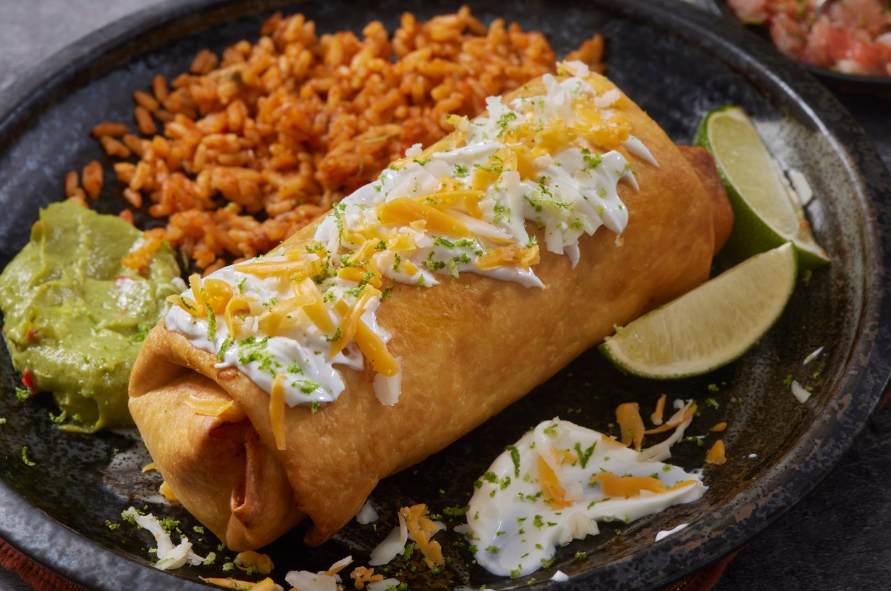 A crispy chimichanga topped with sour cream and shredded cheese, served with Mexican rice, guacamole, and lime wedges on a black plate.