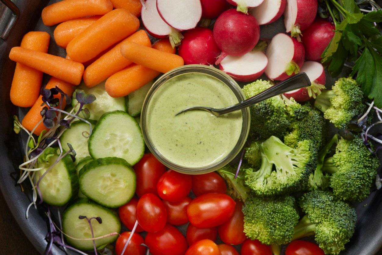 A platter of fresh vegetables, including baby carrots, sliced radishes, cucumber slices, cherry tomatoes, broccoli florets, and greens, arranged around a bowl of green dip with a spoon.
