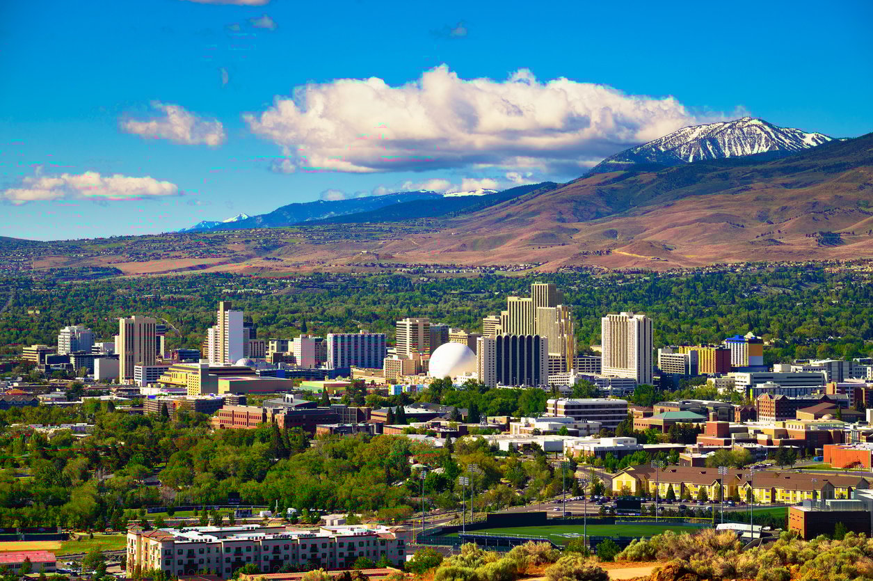A vibrant city skyline with tall buildings set against a backdrop of mountains, some with snow-capped peaks, under a blue sky with scattered clouds and lush green trees in the foreground.
