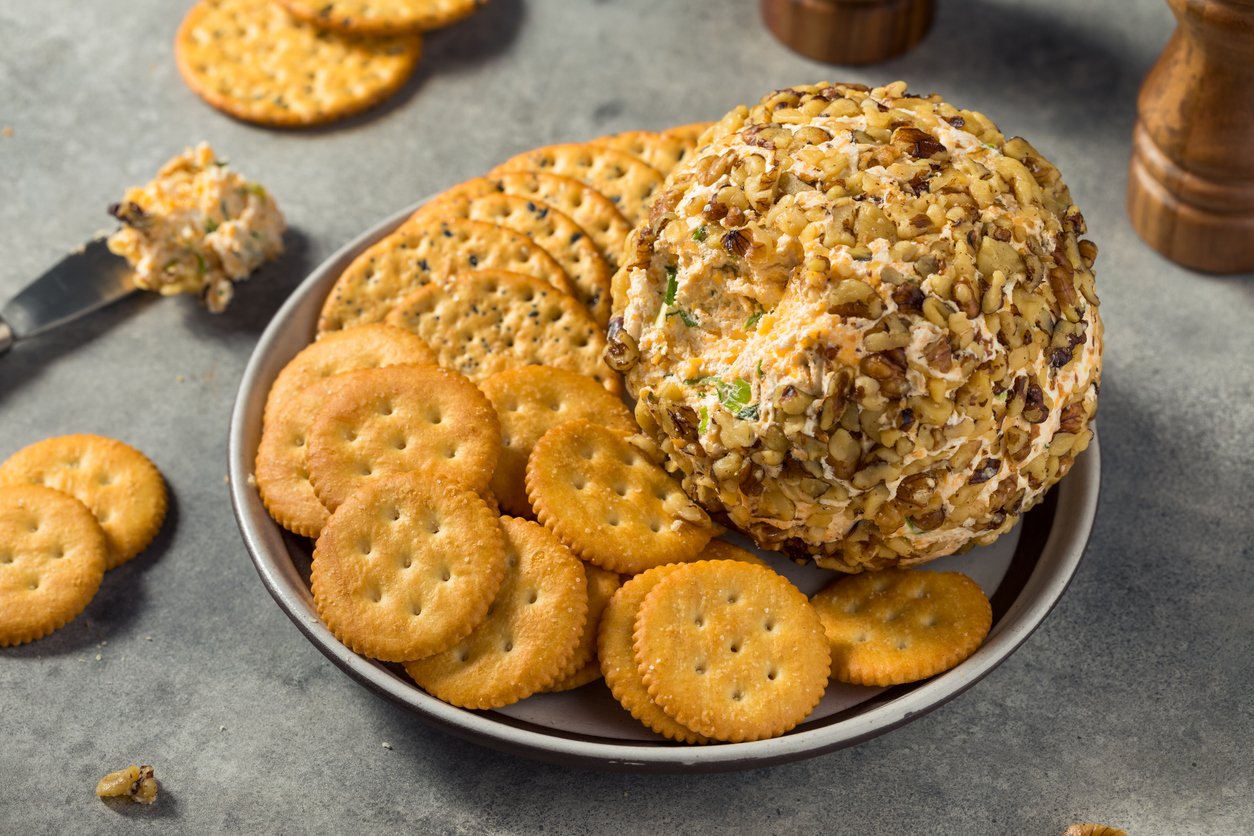 A plate with round crackers and a cheese ball coated in chopped nuts. A knife with spreadable cheese is beside the plate, and some crackers are scattered on the gray surface.