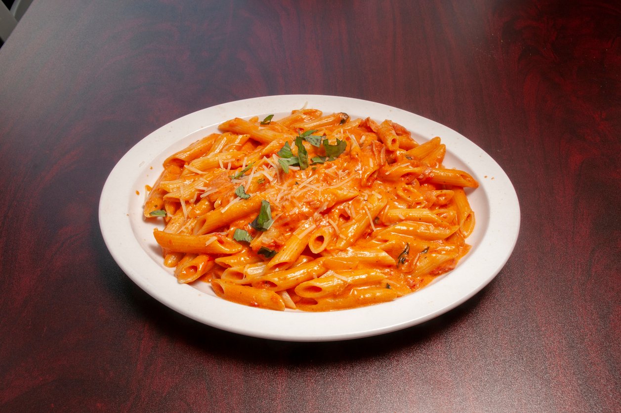 A white plate filled with penne pasta in a creamy tomato sauce, garnished with grated cheese and fresh herbs, sits on a dark wooden table.