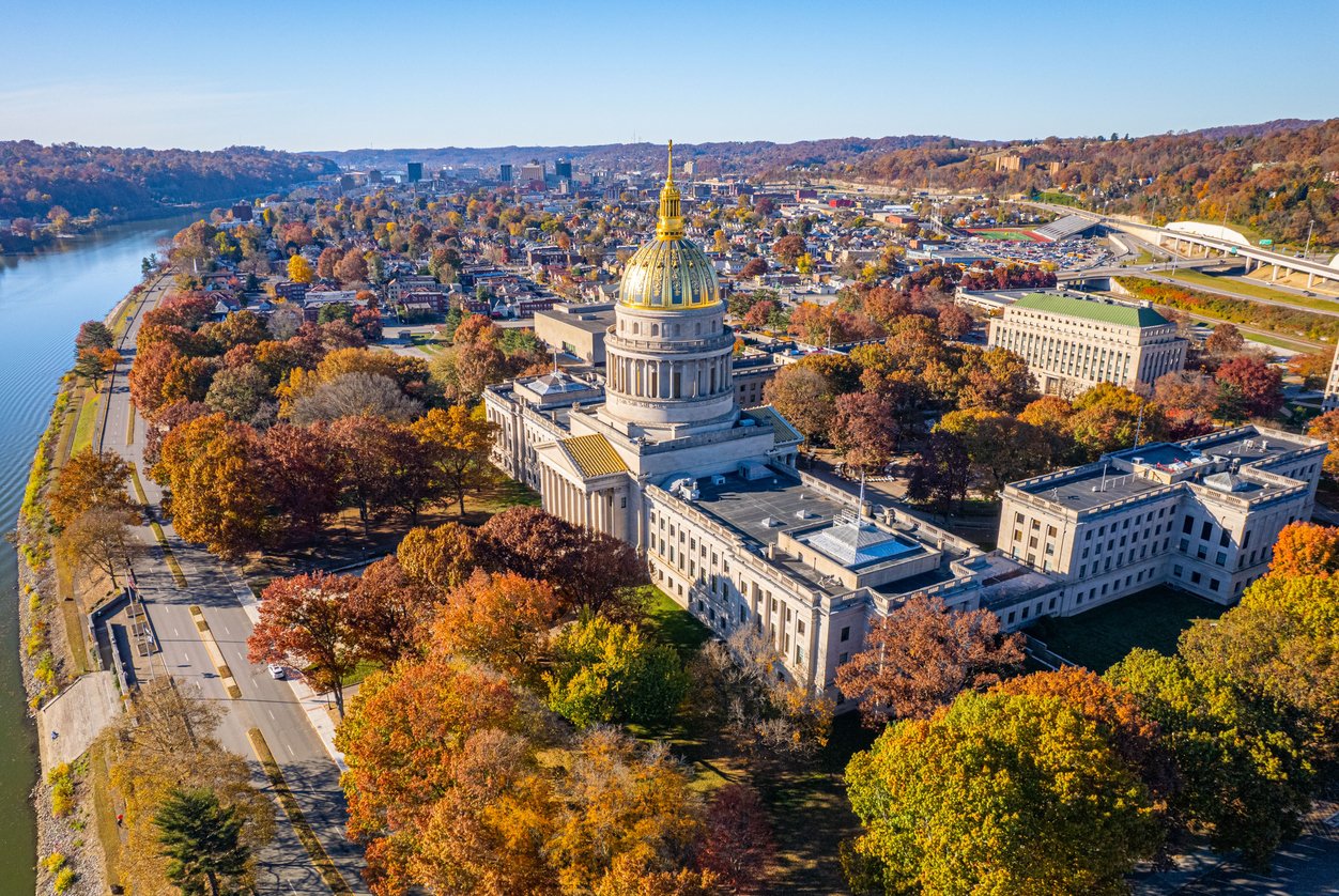 Aerial view of the West Virginia State Capitol building with a gold dome, surrounded by trees with autumn foliage, near a river and cityscape under a clear blue sky.