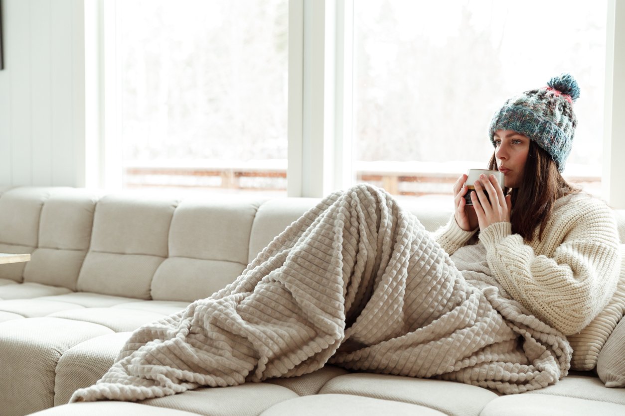 A woman sits on a beige sofa wrapped in a textured blanket, wearing a knit hat and sweater, and sipping from a mug. Large windows behind her reveal a snowy, bright outdoor scene.