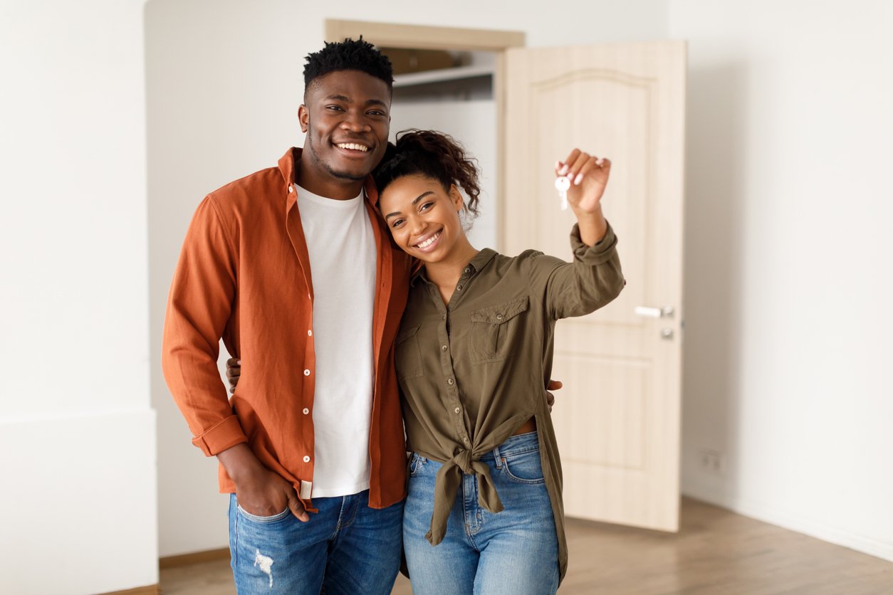 A smiling couple stands in a new home, with one person holding up a key. They appear happy and close, suggesting they just moved in together. An open closet and a door are visible in the background.