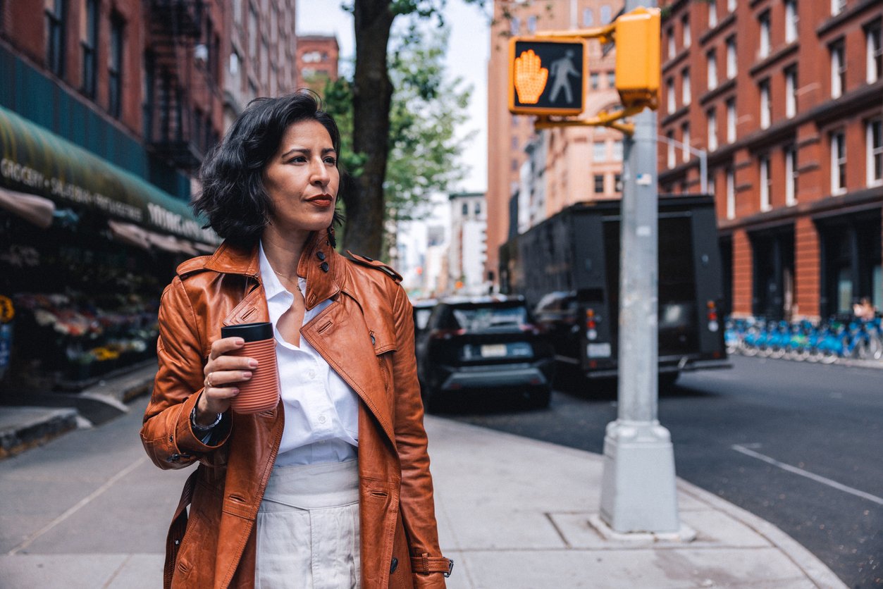 A woman with dark hair, wearing a brown leather jacket and white pants, holds a coffee cup while walking on a city sidewalk. A crosswalk signal and brick buildings are visible in the background.