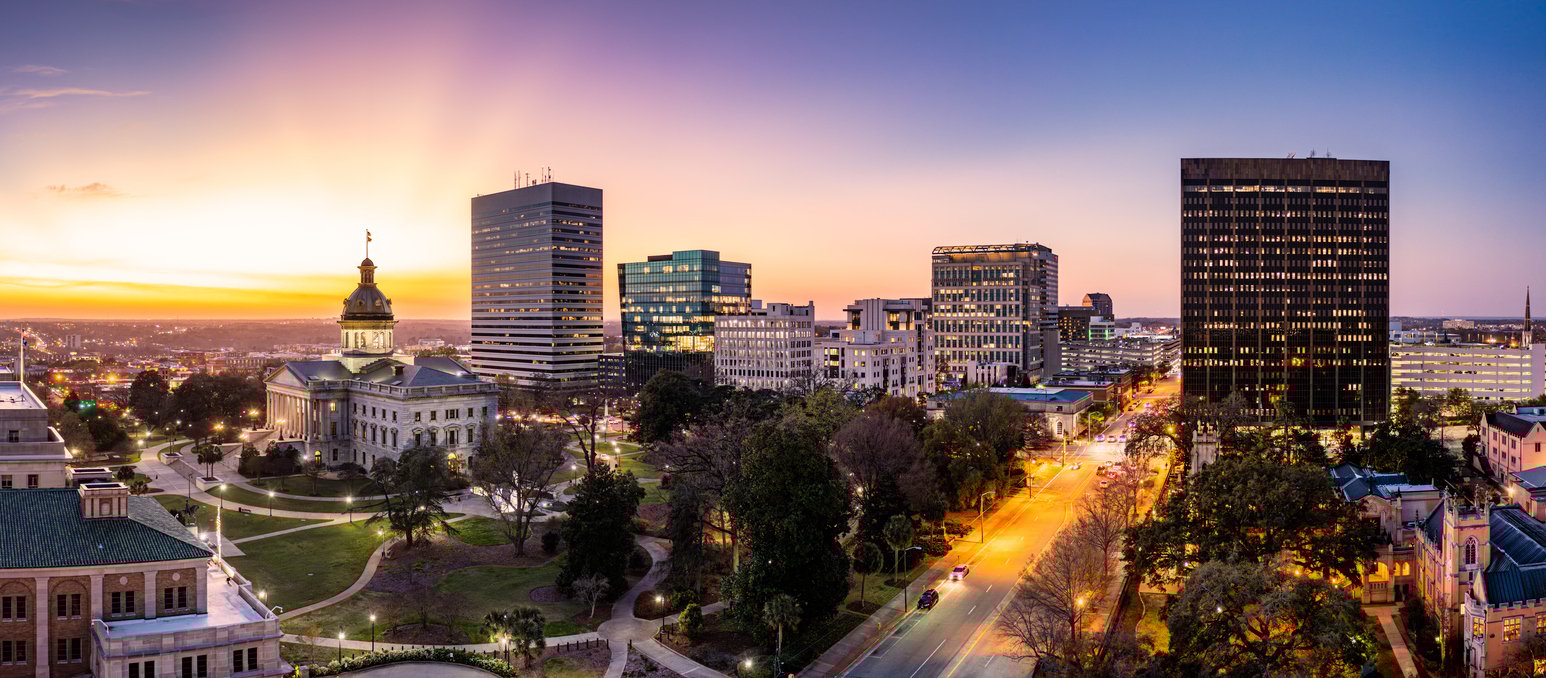 Panoramic view of a Columbia, South Carolina cityscape at sunset featuring a domed capitol building, modern high-rise office buildings, and tree-lined streets with glowing streetlights.