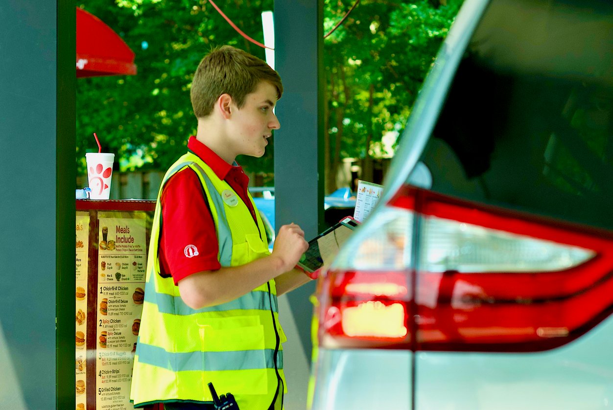 A fast-food worker wearing a red shirt and yellow safety vest takes an order on a tablet beside a car in a drive-thru. A menu and a drink cup are visible in the background.
