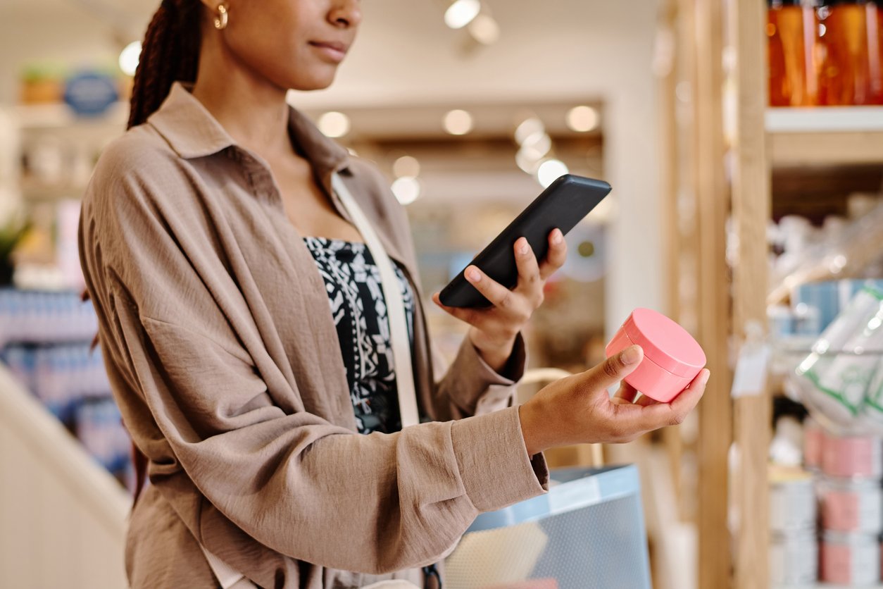 A woman stands in a store holding a pink cosmetic jar in one hand and a smartphone in the other, appearing to scan or research the product. She is wearing a light brown jacket and patterned dress. Shelves are visible in the background.