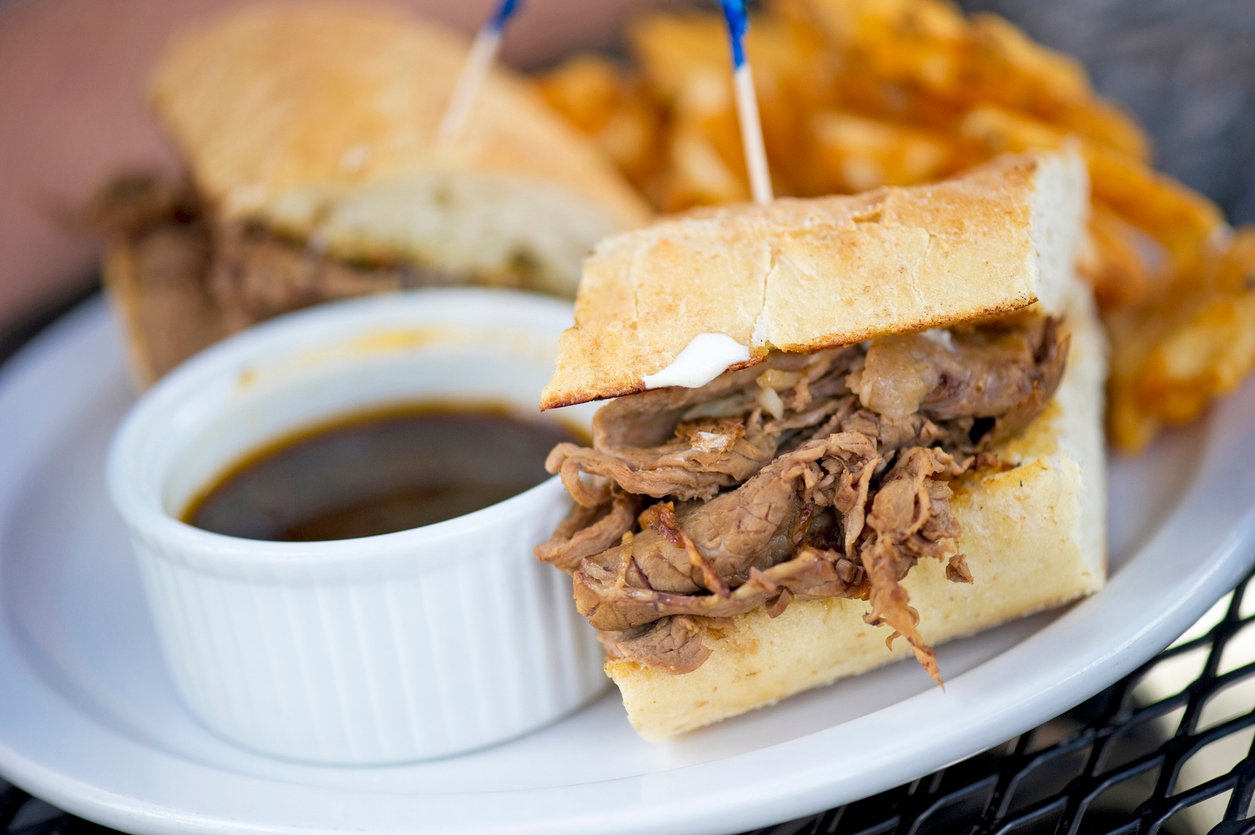 A French dip sandwich filled with sliced roast beef on a white plate, served with a small bowl of au jus and a side of waffle fries.