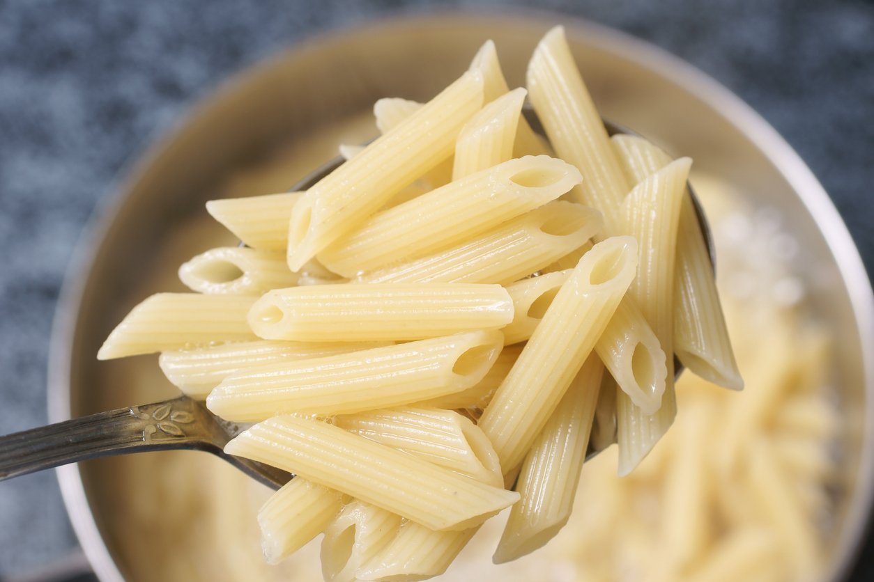A close-up of a spoon holding cooked penne pasta above a pot filled with more pasta. The penne is plain and lightly glistening with steam visible, indicating it is freshly boiled.