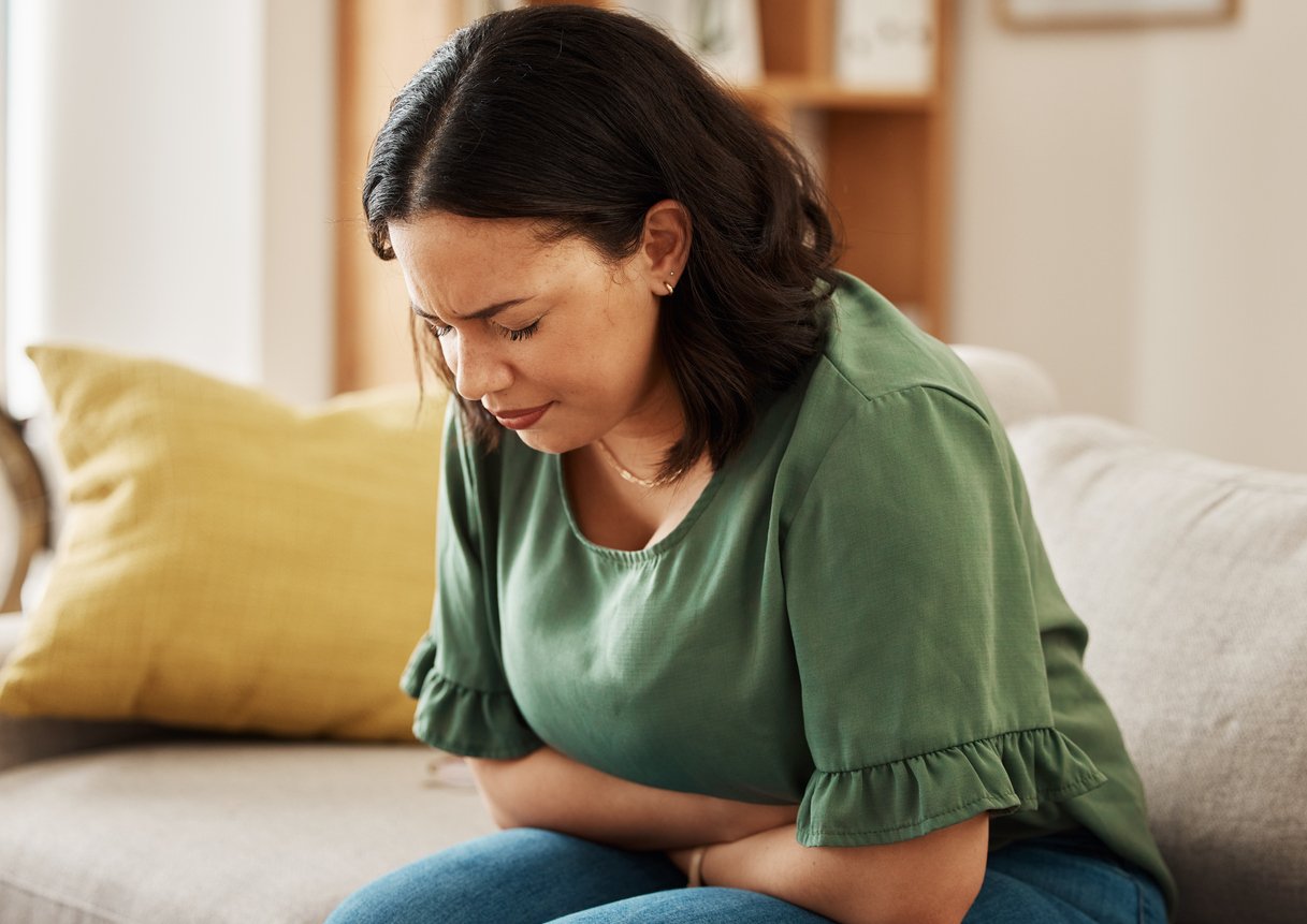 A woman sits on a couch, leaning forward and holding her stomach with both hands, appearing to be in pain or discomfort.