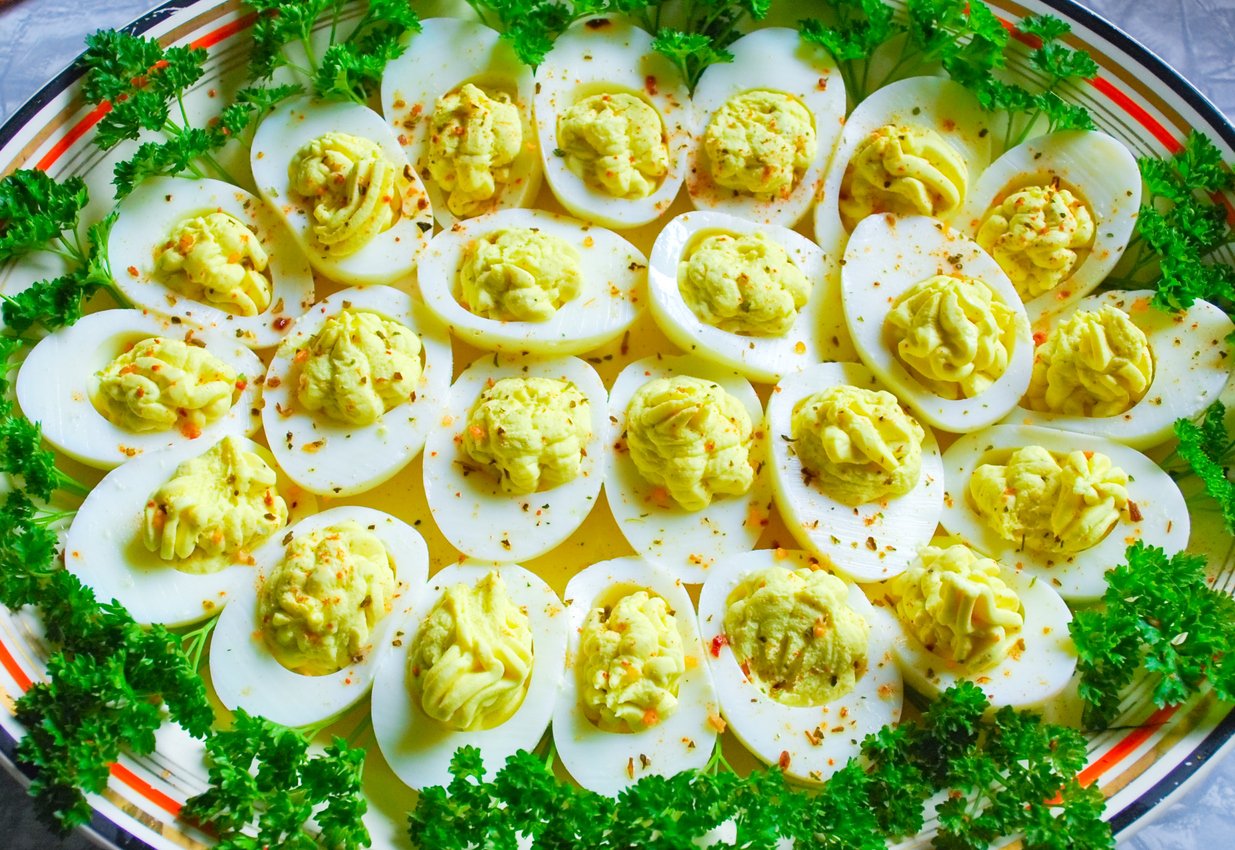 A platter of deviled eggs garnished with parsley. The eggs are halved, filled with a creamy yellow yolk mixture, and sprinkled with spices. Parsley is arranged around the edges of the plate for decoration.