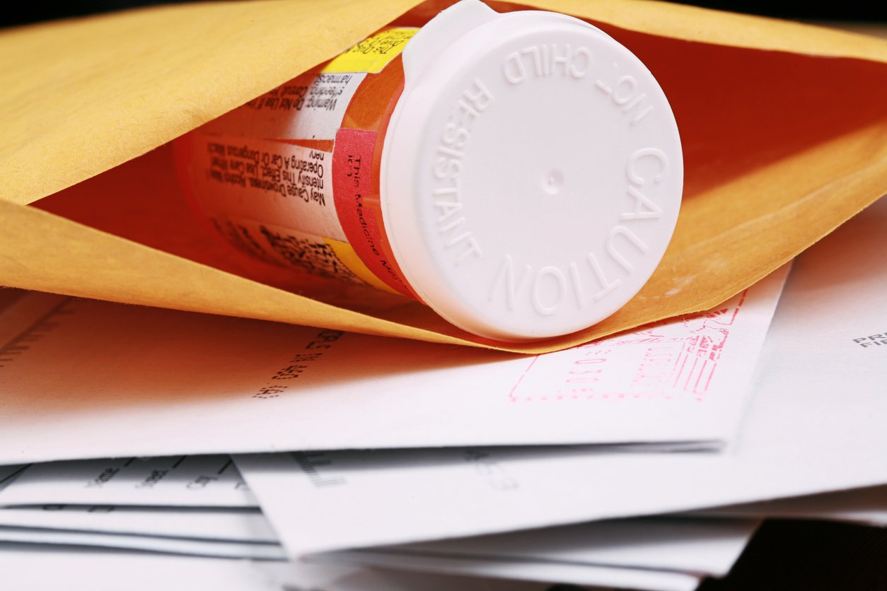 A prescription pill bottle partially visible inside a yellow envelope, resting on a stack of mail and documents.