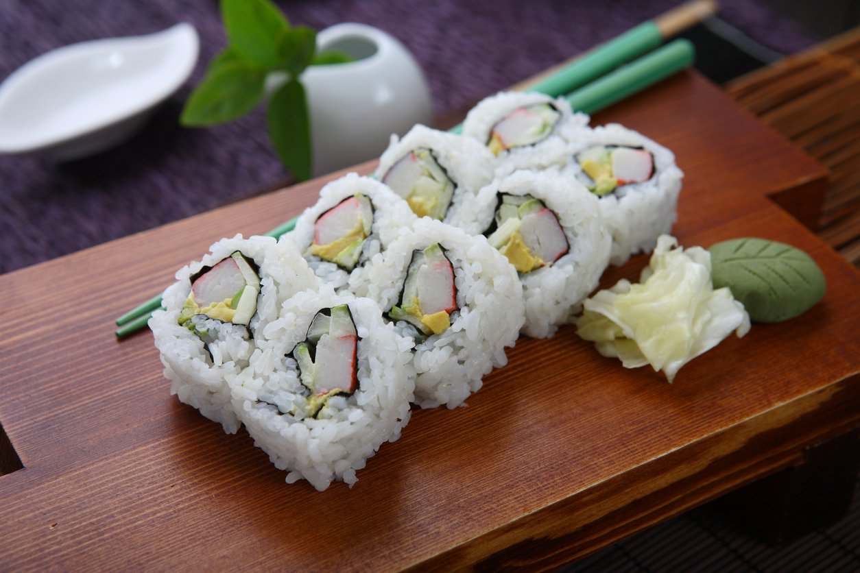 Six California sushi rolls with crab, avocado, and cucumber are arranged on a wooden tray with pickled ginger, wasabi, and green chopsticks, with a leaf and small dishes in the background.