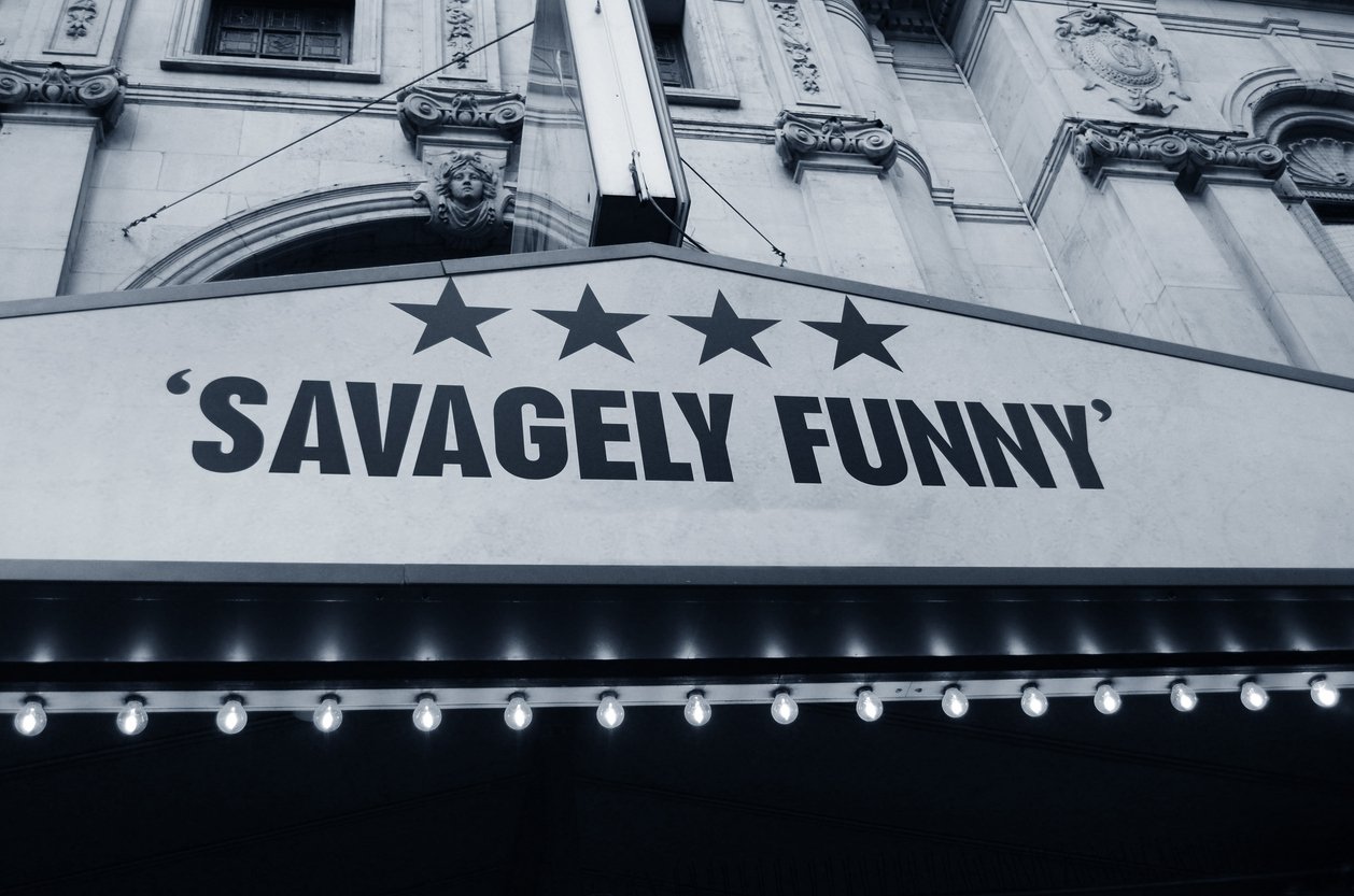 A theater marquee reads “SAVAGELY FUNNY” in bold letters, with four stars above the text. The sign is lit with small bulbs below, and a decorative stone building is visible in the background.