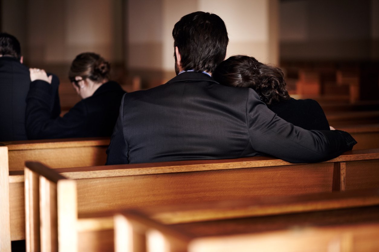 Four people sitting in wooden pews; two in the foreground, with one resting their head on the other's shoulder, both dressed formally, in a somber or reflective setting, possibly a church or memorial service.