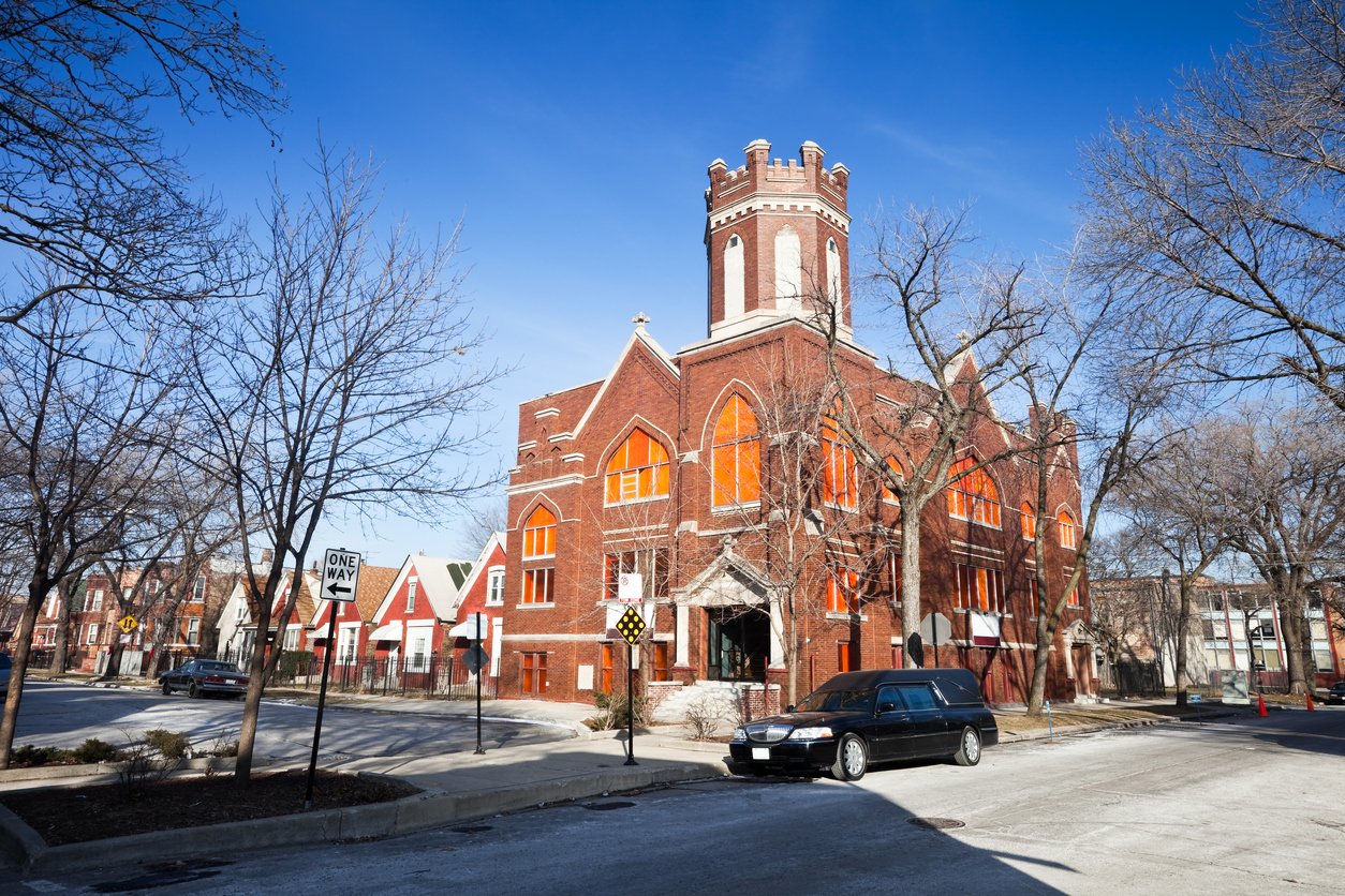 A historic red brick church with large orange windows and a castle-like tower stands on a street corner with leafless trees, parked cars, and houses nearby under a clear blue sky.