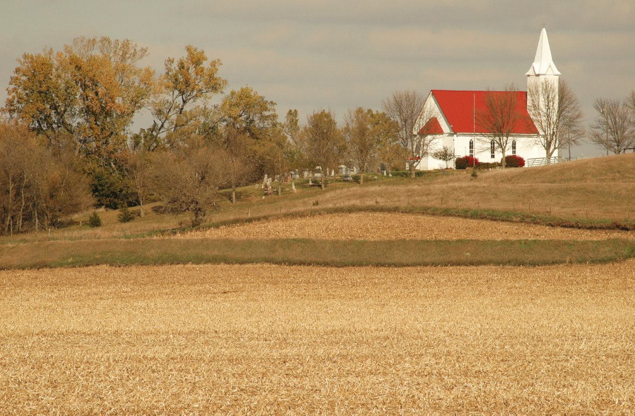 A white church with a red roof stands on a hilltop surrounded by autumn trees and a graveyard, overlooking a harvested cornfield under a partly cloudy sky.