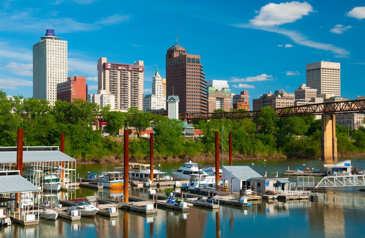 Boats docked at a marina on a river with a city skyline in the background, green trees along the shore, and a bridge crossing over the water under a blue sky with scattered clouds.