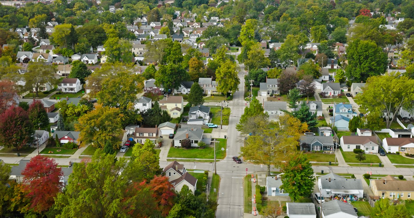 Aerial view of a suburban neighborhood with tree-lined streets, single-family homes, and green lawns. The trees display a mix of green and some autumn colors, and the houses are evenly spaced along the roads.