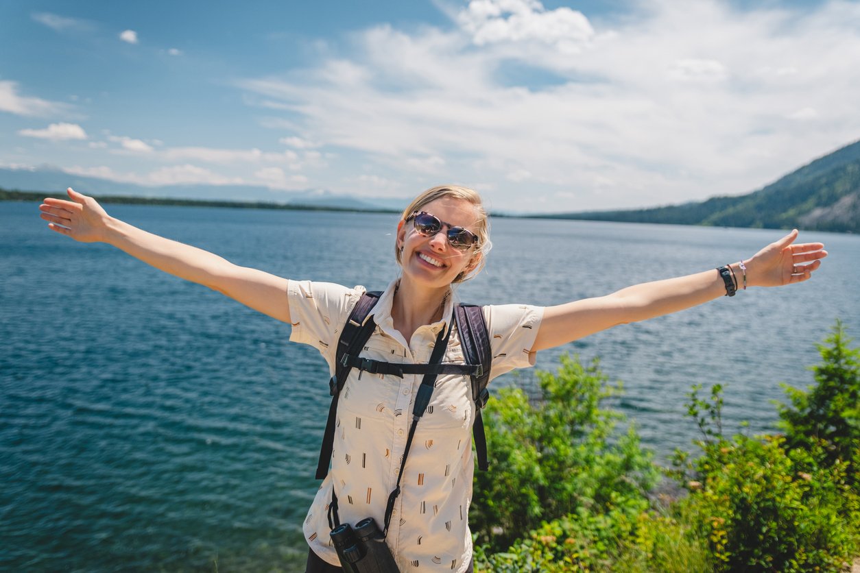A smiling person with sunglasses and a backpack stands by a lake, arms outstretched, with green foliage and mountains in the background on a sunny day.