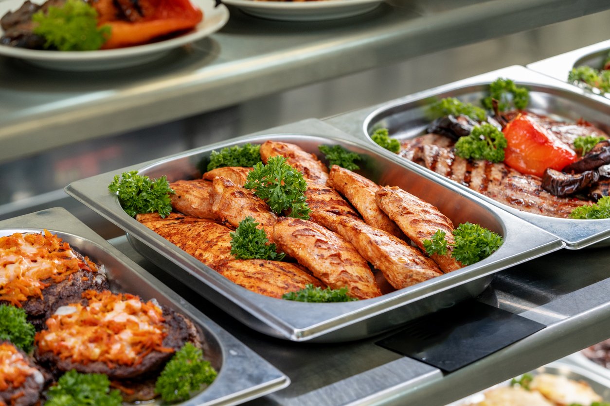 Metal trays filled with grilled chicken breasts garnished with parsley, along with roasted vegetables, are displayed on a serving counter in a cafeteria or buffet setting.