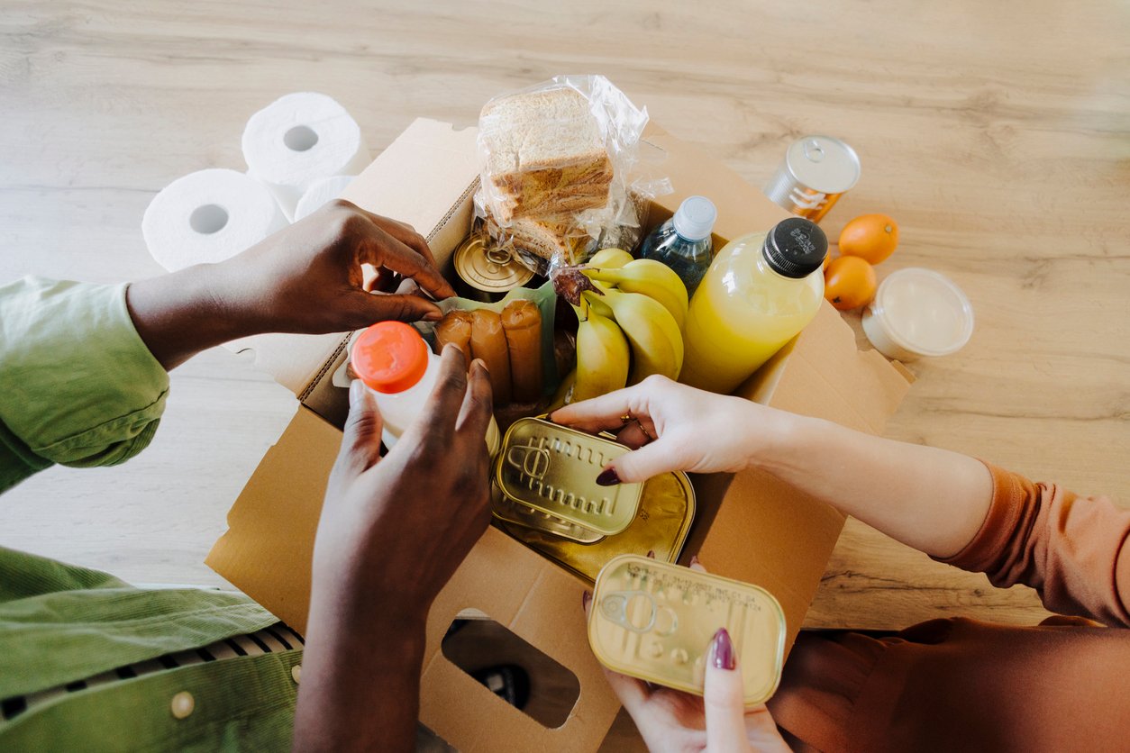 Two people are unpacking a box of groceries containing canned food, bananas, orange juice, eggs, bread, tomatoes, and rolls of toilet paper on a wooden table.