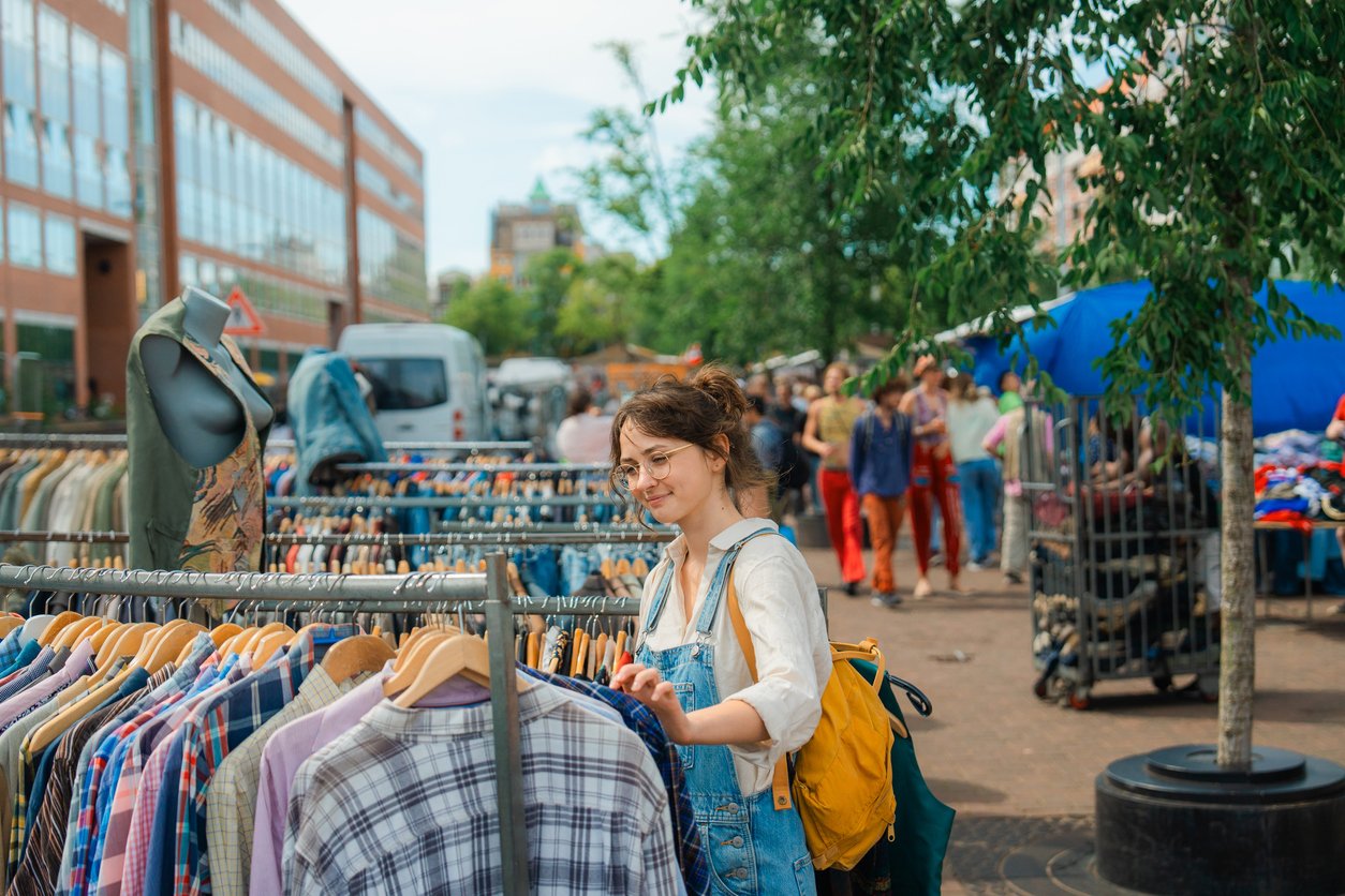 A young woman with glasses and a yellow backpack browses clothes on racks at an outdoor market, surrounded by other shoppers and colorful merchandise on a sunny day.