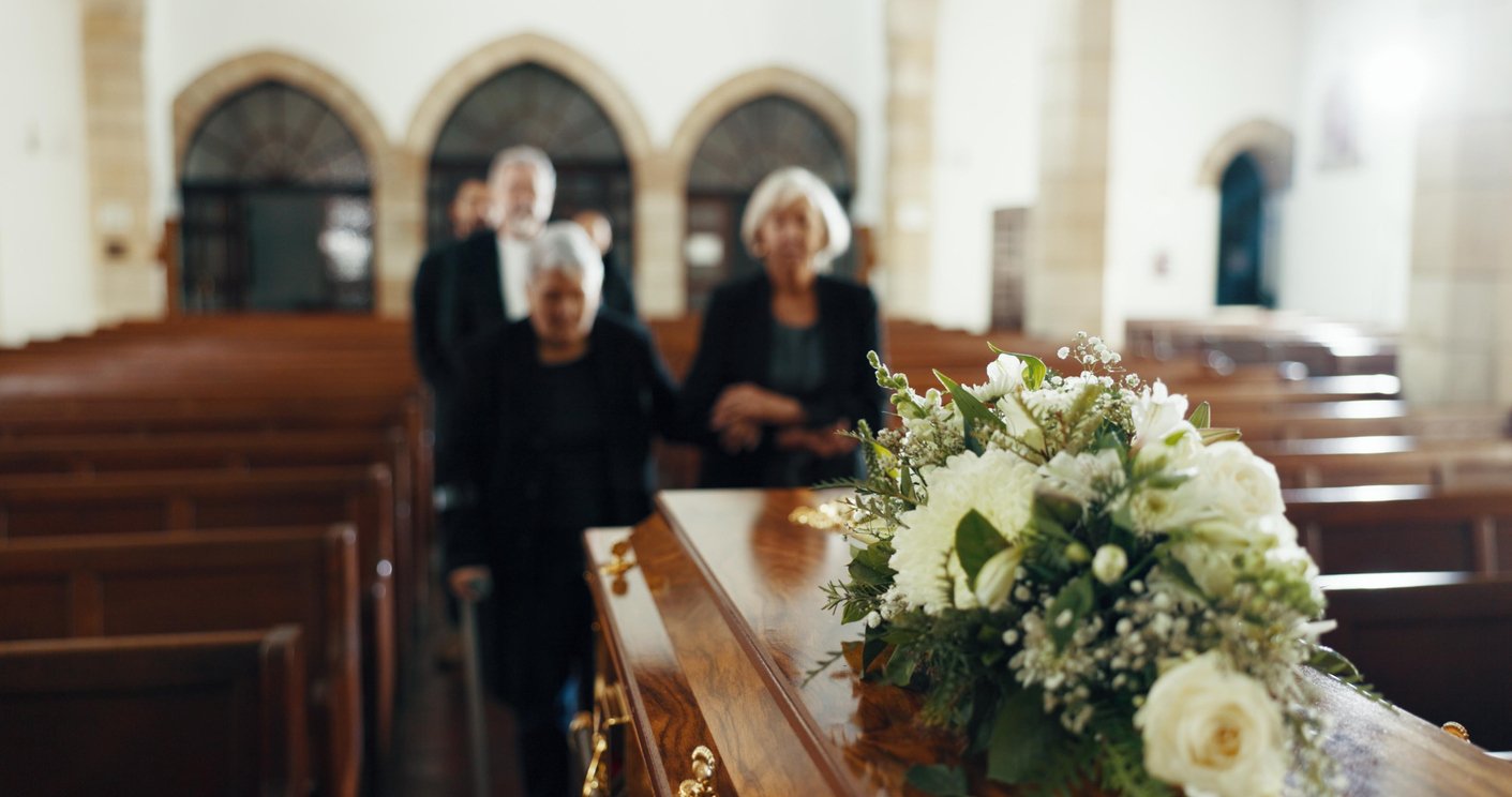 A close-up of a wooden casket with white flowers on top in a church. In the background, three mourners dressed in black walk down the aisle toward the casket. The scene is solemn and softly lit.