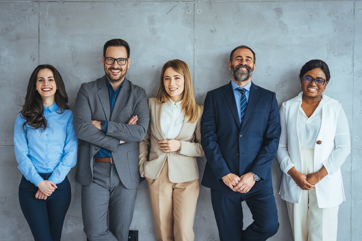 Five professionally dressed people stand in a row against a gray wall, smiling confidently at the camera. The group includes three women and two men in business attire.