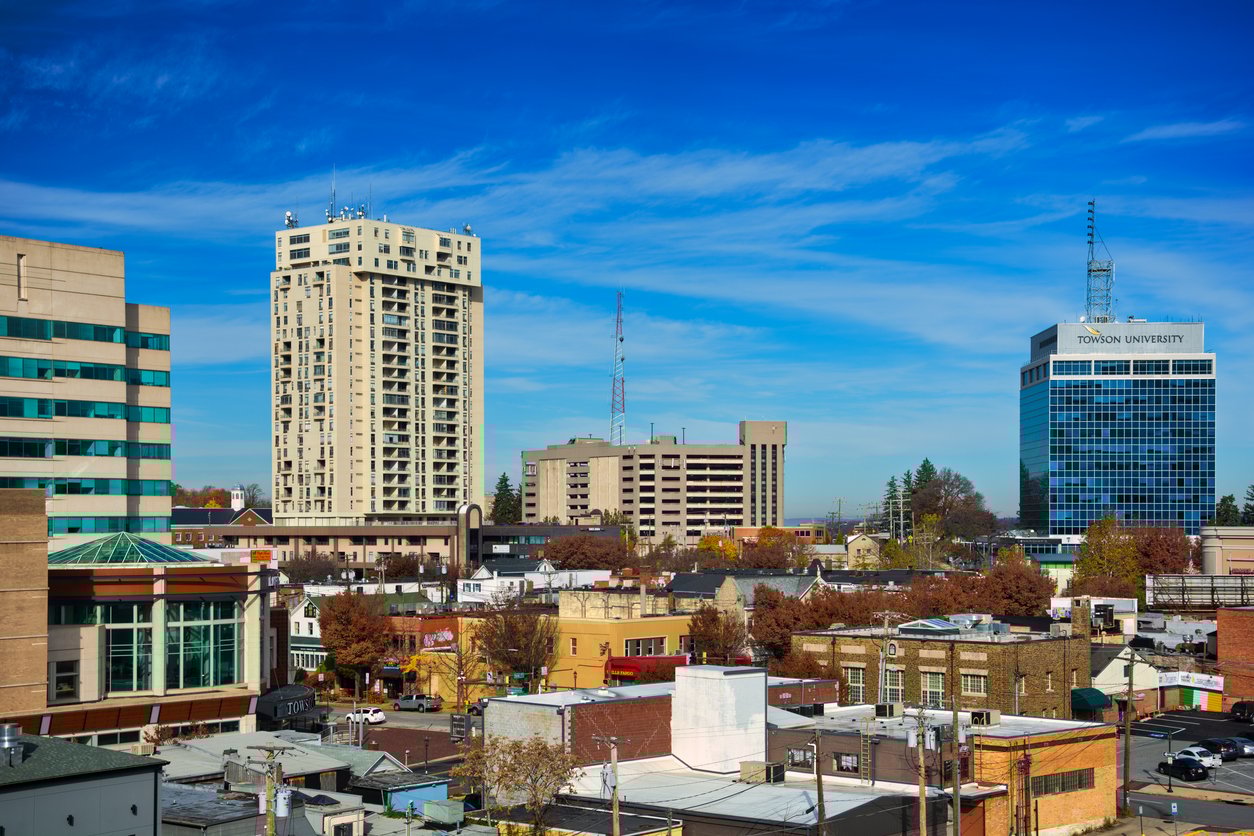 View of a mid-sized city with low-rise buildings in the foreground, taller office and apartment buildings in the background, autumn trees, and a clear blue sky overhead. One building displays "Toronto University.