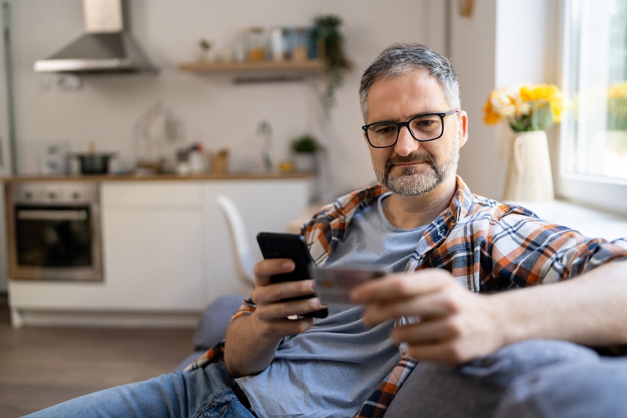 A man with glasses sits on a couch in a bright living room, holding a smartphone in one hand and a credit card in the other, appearing focused as he looks at the card.