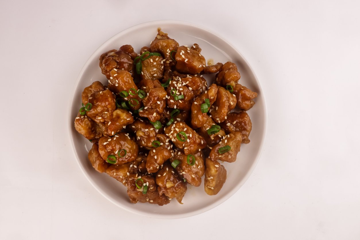 A white plate filled with pieces of glazed fried chicken garnished with sliced green onions and sesame seeds, set against a plain white background.