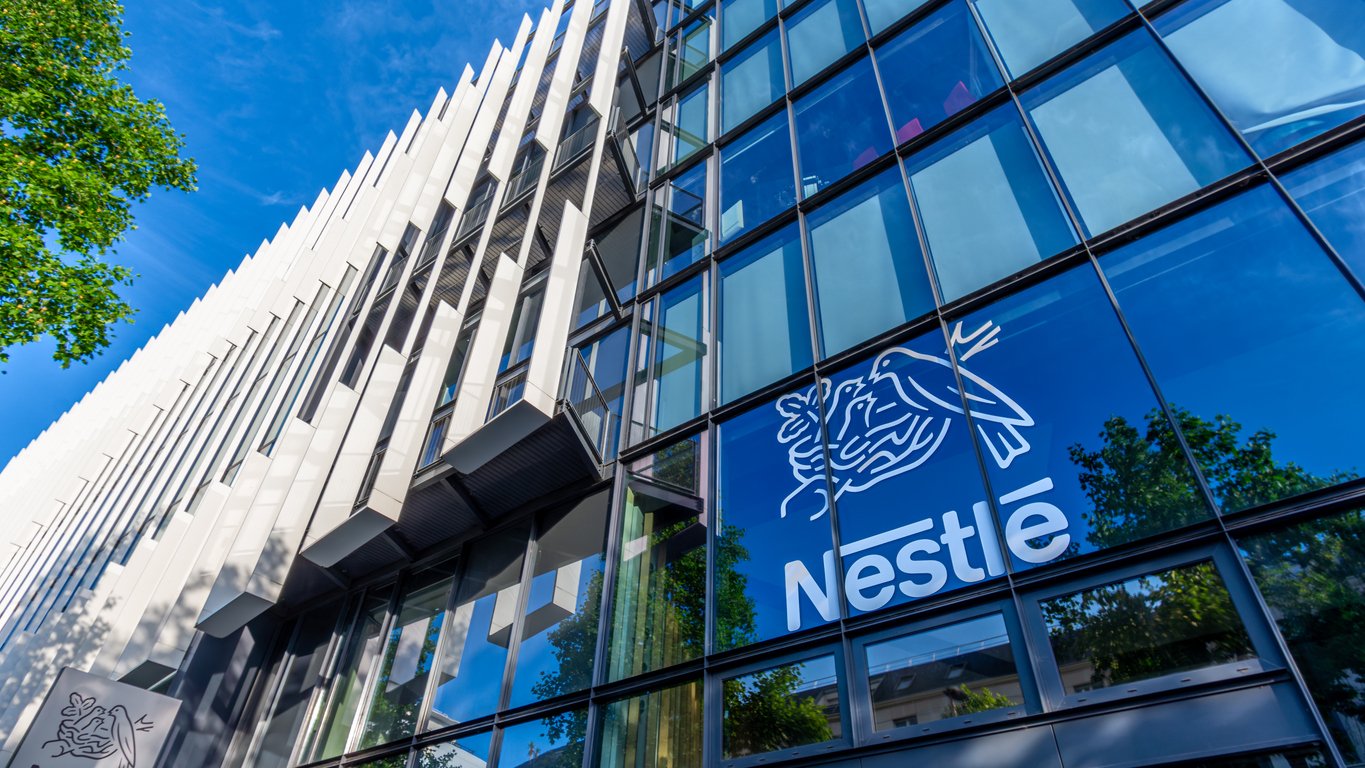 Modern glass building with the Nestlé logo, featuring a bird feeding its chicks, prominently displayed on the lower right. Reflections of trees and a blue sky are visible in the windows.
