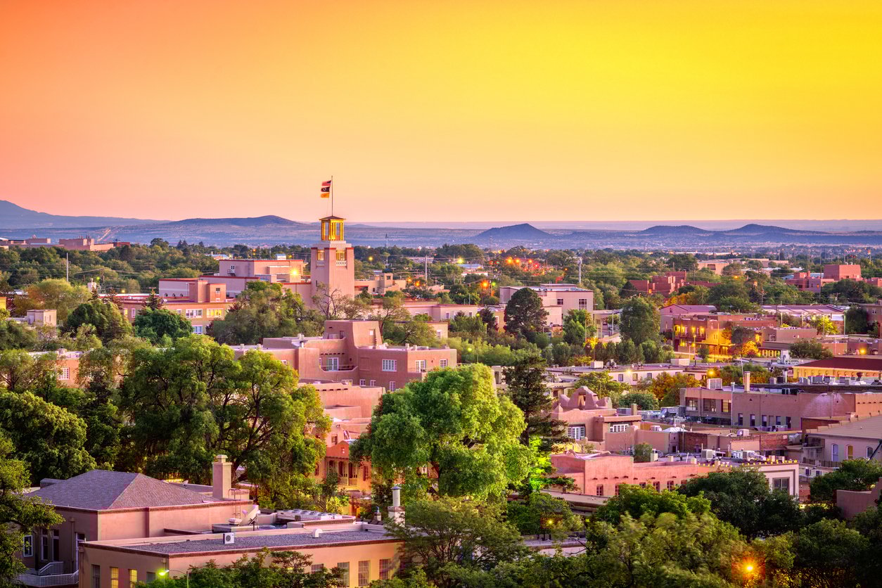 Santa Fe, New Mexico skyline at sunset with adobe-style buildings, lush green trees, and a prominent building flying a flag, set against distant hills and a vibrant yellow-orange sky.
