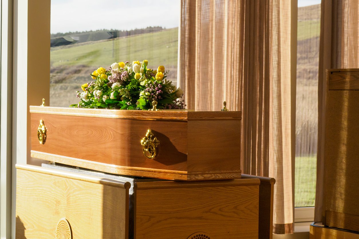 A wooden coffin adorned with a flower arrangement sits on a stand in a sunlit room, with sheer curtains and a view of grassy hills through the window in the background.